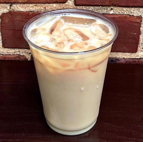 A clear plastic cup filled with iced coffee, featuring cream swirling into the dark liquid, set against a brick background.
