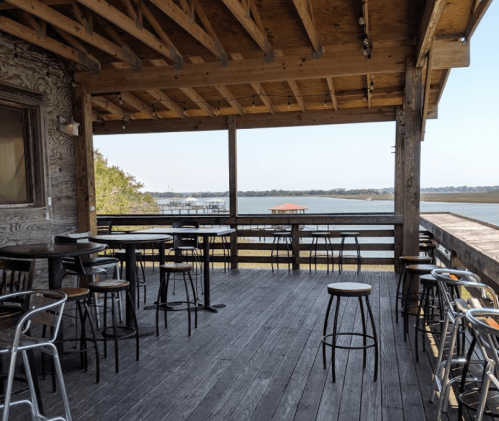 A wooden deck with empty bar stools overlooks a calm water view and distant structures under a clear sky.