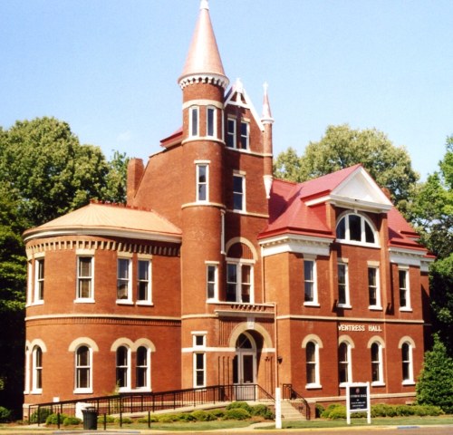 Historic brick building with a turret and pointed roof, surrounded by trees and a clear blue sky.