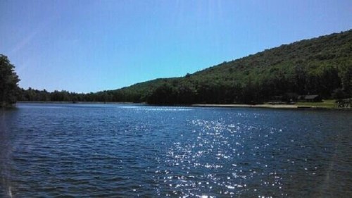 A serene lake surrounded by lush green hills under a clear blue sky, with sunlight sparkling on the water's surface.