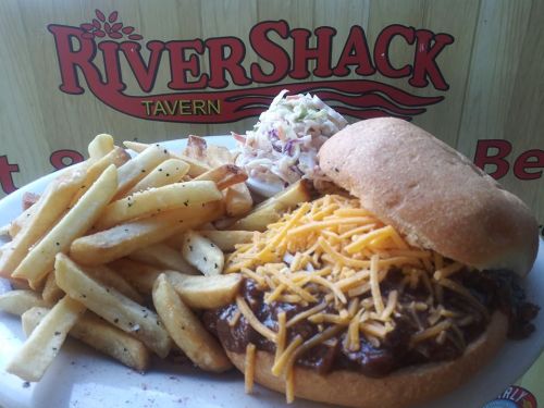 A plate featuring a sandwich with chili and cheese, served with fries and coleslaw, against a RiverShack Tavern sign.