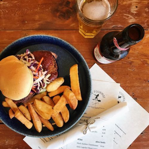 A plate with a burger topped with coleslaw, golden fries, a glass of beer, and menus on a wooden table.