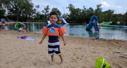 A smiling boy in a life jacket stands on the sandy beach by a pool, with inflatable toys and other children in the background.
