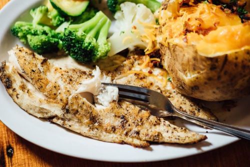 A plate with grilled fish, steamed broccoli, cauliflower, and a baked potato topped with cheese and bacon.