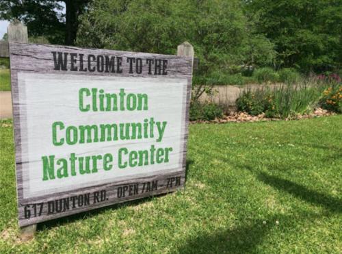 Sign welcoming visitors to the Clinton Community Nature Center, with hours and address details. Green grass and plants in background.
