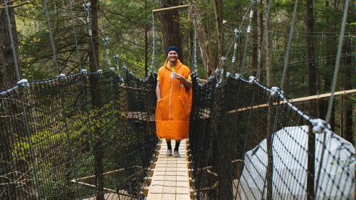 A person in an orange coat stands on a wooden bridge in a forest, holding a cup and smiling.