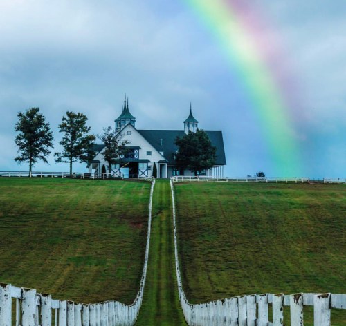 A picturesque white barn with spires, set on a green hill under a rainbow and cloudy sky, framed by a white fence.