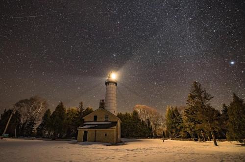A lighthouse shines brightly under a starry night sky, surrounded by snow-covered ground and trees.