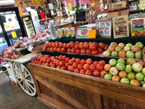 A wooden cart filled with red and green apples and tomatoes in a market setting, with signs and products in the background.