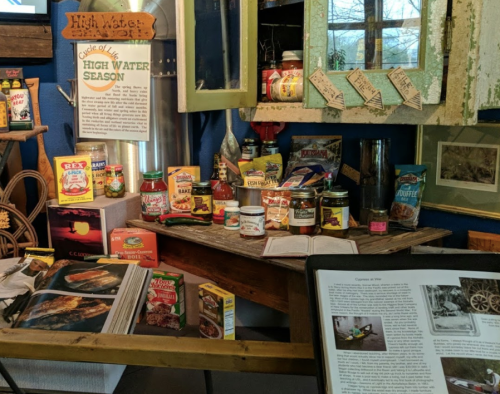 Display of vintage food cans and jars on a wooden table, with informational signs and a book in a museum setting.