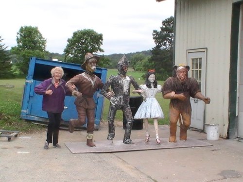 A woman stands next to large metal sculptures of characters from "The Wizard of Oz" in an outdoor setting.