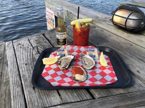 A tray with oysters, lemon wedges, a beer, and a cocktail sits on a wooden dock by the water.