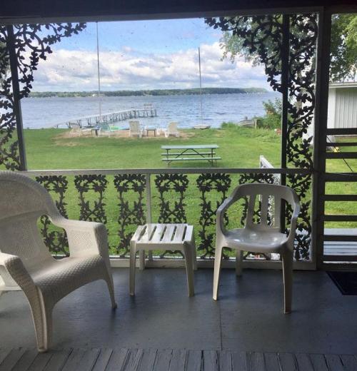 A screened porch with two white chairs overlooks a lake and grassy area with picnic tables and a dock in the distance.