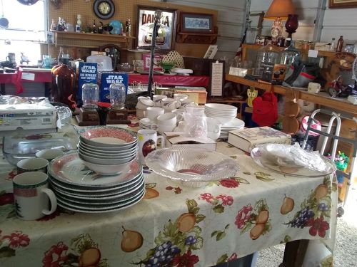 A cluttered table in a garage sale featuring various dishes, cups, and collectibles, including Star Wars items.