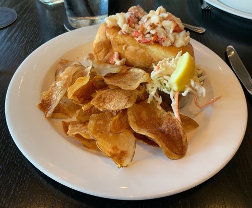 A lobster roll topped with lobster meat, served with crispy potato chips and a side of coleslaw on a white plate.