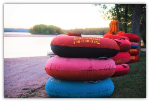 Colorful inner tubes stacked by a riverbank at sunset, with a resort's contact number visible on one tube.