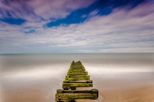 A weathered wooden pier extends into a calm sea under a cloudy sky, with soft waves lapping at the shore.
