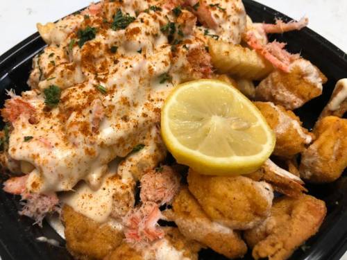 A black container filled with shrimp, fried seafood, and a lemon slice, topped with a creamy sauce and seasoning.