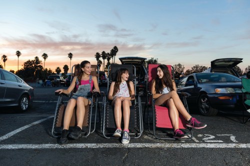 Three girls sit in reclining chairs in a parking lot at sunset, chatting and enjoying the evening.