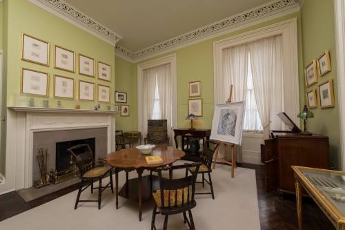 A cozy room with green walls, a round table, chairs, artwork on display, and a fireplace. Natural light from windows.