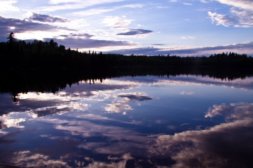 A serene lake at sunset, reflecting clouds and trees under a colorful sky.