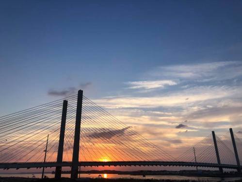 Silhouette of a cable-stayed bridge against a vibrant sunset sky with clouds and water below.