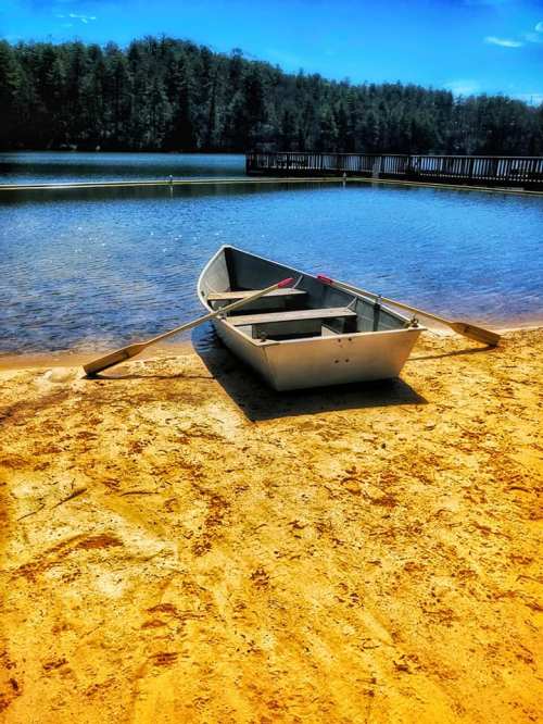 A small boat on a sandy shore by a calm lake, surrounded by trees and a wooden dock in the background.