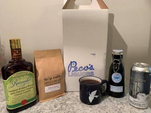 A bottle of bourbon, coffee bag, a mug, a soda bottle, and a beer can arranged next to a white takeout bag.