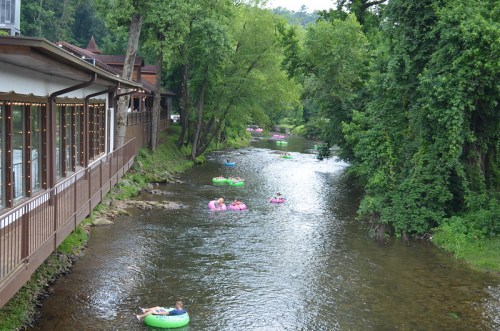 A serene river scene with colorful inner tubes floating, surrounded by lush greenery and a wooden building nearby.