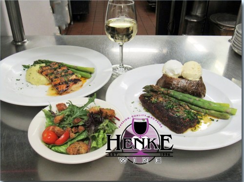 A gourmet meal featuring salmon, steak, baked potato, asparagus, and a fresh salad, with a glass of white wine.