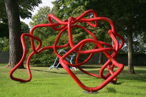 A large, abstract red sculpture made of intertwined tubes, set in a grassy park with trees in the background.