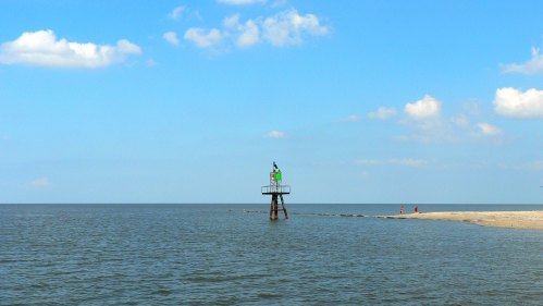 A green buoy stands in calm waters under a blue sky with scattered clouds, near a sandy shore.