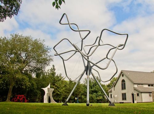 A modern outdoor sculpture with abstract shapes stands in a grassy area, alongside another artistic structure and a building.