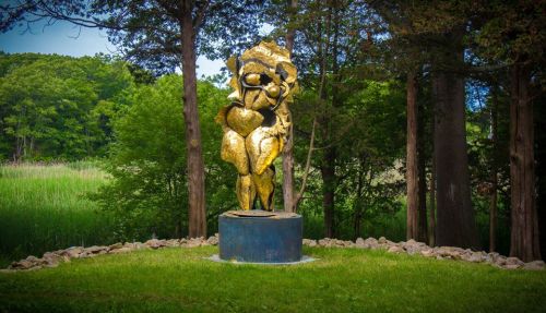 A golden abstract sculpture stands on a circular base, surrounded by trees and greenery in a serene outdoor setting.
