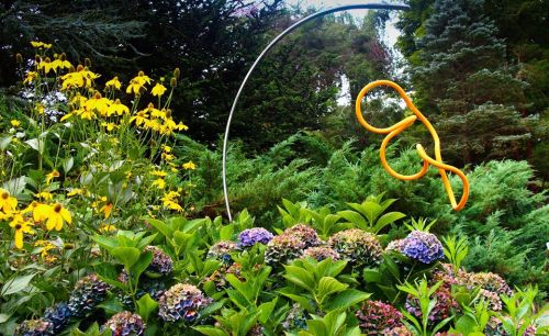 A vibrant garden scene featuring yellow flowers, lush greenery, and an abstract orange sculpture amidst colorful hydrangeas.