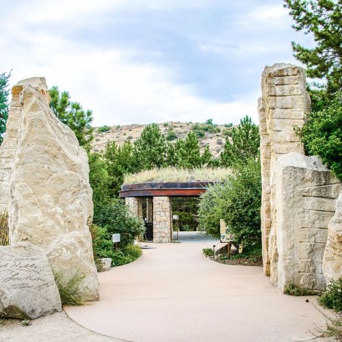 A stone archway leads into a lush garden, surrounded by greenery and hills under a cloudy sky.