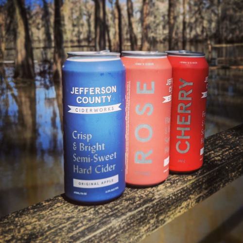Three cans of cider on a wooden railing, with flavors labeled: Original Apple, Rose, and Cherry, set against a natural backdrop.