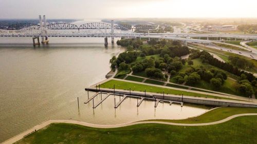 Aerial view of a river with a bridge, green parkland, and a marina under a hazy sky at sunset.