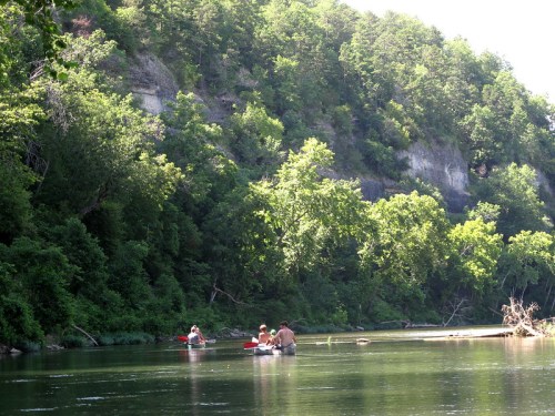 Two people kayak on a calm river surrounded by lush green trees and steep cliffs in the background.