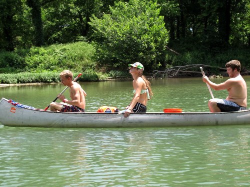 Three young people canoeing on a river, enjoying a sunny day, surrounded by lush greenery.