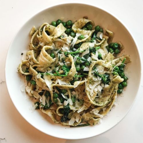 A bowl of pasta with pesto, peas, and shaved cheese, garnished with black pepper on a white surface.