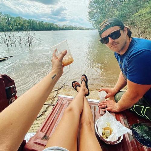 A person relaxes by a river, holding a drink, while another sits nearby, both enjoying a sunny day outdoors.