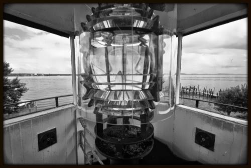 A close-up of a lighthouse lens with a view of the water and pier in the background, captured in black and white.