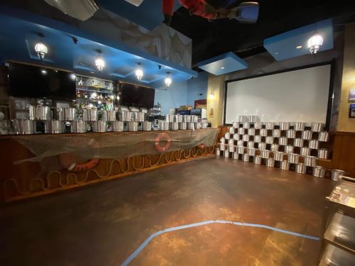 A dimly lit bar area with a counter, shelves of metal containers, and a large screen in the background.