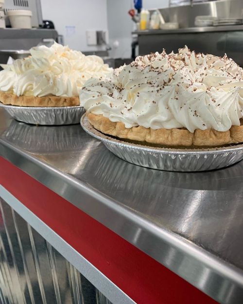 Two cream-topped pies on a stainless steel counter, one with chocolate shavings and the other plain.