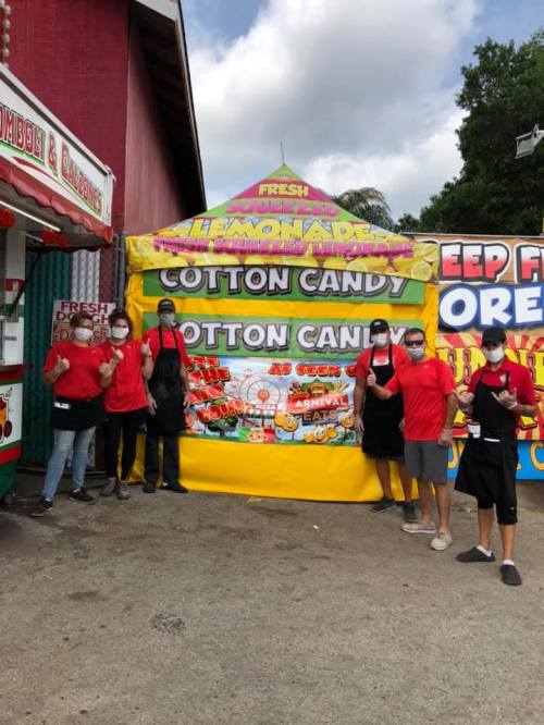 A group of six people in red shirts and masks stand in front of a colorful booth selling cotton candy at a fair.