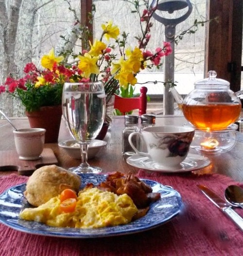 A cozy breakfast table with scrambled eggs, bacon, a biscuit, tea, and fresh flowers in a rustic setting.