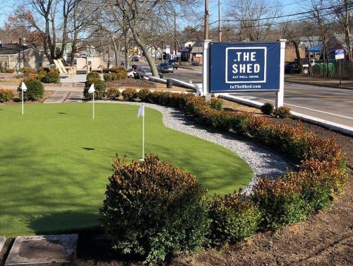 A green putting green with flags, bordered by shrubs, next to a sign for "The Shed" restaurant on a busy street.