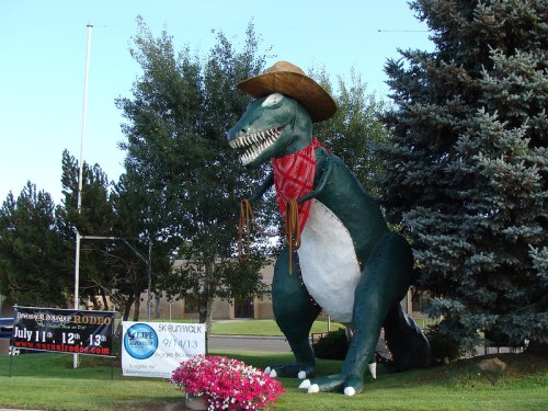 A large dinosaur statue wearing a sombrero and scarf, standing next to a sign and colorful flowers.