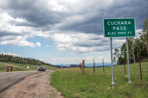 A road sign for Cuchara Pass at 9995 feet elevation, with a car parked nearby and a scenic landscape in the background.